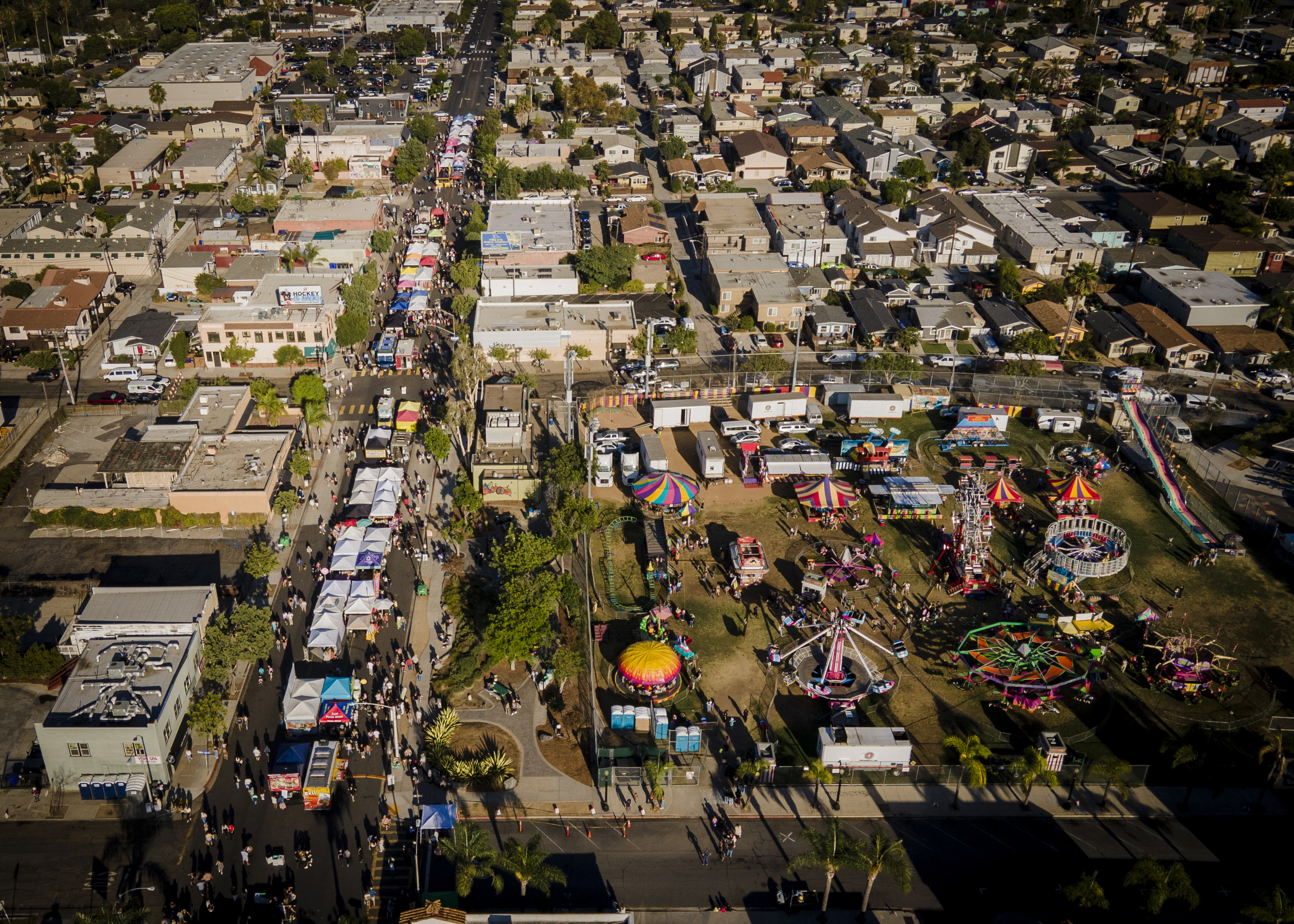 Aerial view of a lively street fair with tents, food trucks, and carnival rides on Adams Avenue.